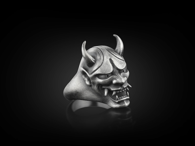 May include: A silver ring with a detailed oni mask design. The mask has horns, sharp teeth, and a fierce expression.
