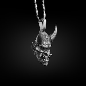 May include: A silver pendant necklace featuring a detailed, three-dimensional Oni mask with horns. The mask has a fierce expression with sharp teeth and a textured surface.