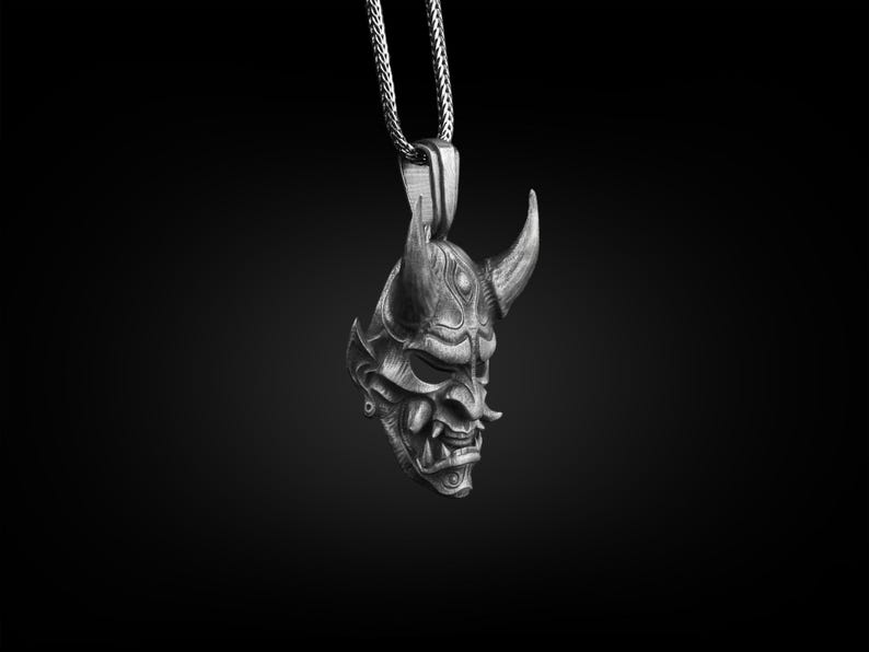 May include: A silver pendant necklace featuring a detailed, three-dimensional Oni mask with horns. The mask has a fierce expression with sharp teeth and a textured surface.