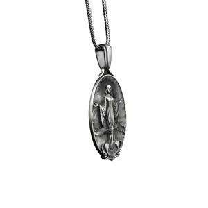 Puede incluir: Collar con colgante de plata con un dije ovalado con una imagen detallada de una mujer con una t&uacute;nica fluida. El dije tiene una superficie texturizada y detalles intrincados.