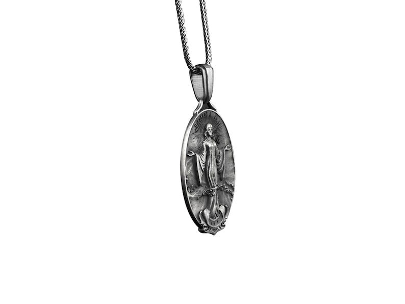 Puede incluir: Collar con colgante de plata con un dije ovalado con una imagen detallada de una mujer con una t&uacute;nica fluida. El dije tiene una superficie texturizada y detalles intrincados.