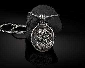 Puede incluir: Un collar de plata con un colgante redondo que presenta un relieve detallado de Jesucristo con una corona de espinas. El colgante está sobre una superficie de roca negra.