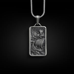 May include: A silver pendant necklace featuring a rectangular charm with a detailed engraving of a mythical sea god holding a trident and riding a ship.