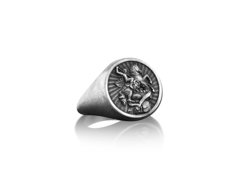 Puede incluir: Un anillo de sello de plata con un dise&ntilde;o detallado de un caballero matando a un drag&oacute;n.