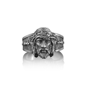 May include: A silver ring with a detailed depiction of Jesus' face, wearing a crown of thorns. The ring features a cross design on the band.