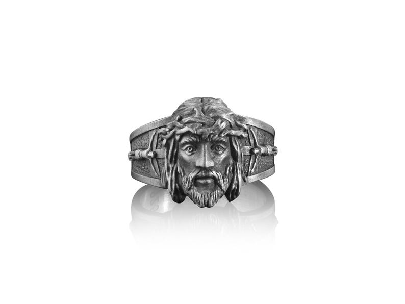May include: A silver ring with a detailed depiction of Jesus' face, wearing a crown of thorns. The ring features a cross design on the band.
