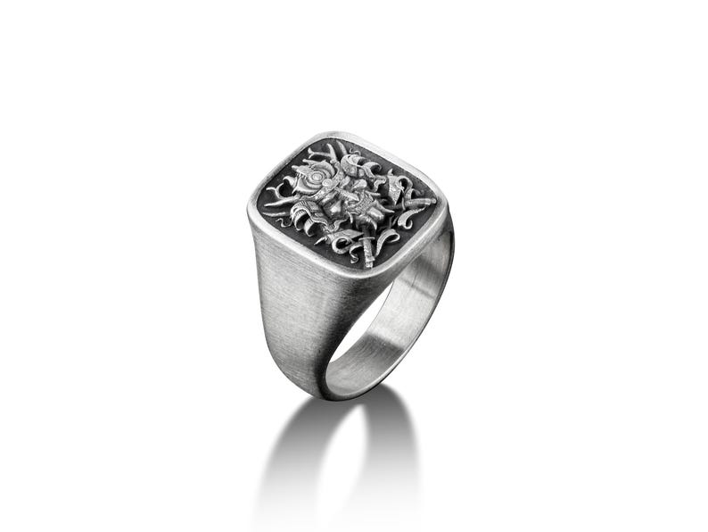 May include: A silver signet ring with a square face featuring a detailed engraved design of a mythical creature with wings and a crown.