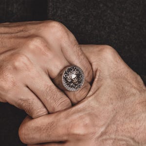 Puede incluir: Un anillo de plata con un diseño detallado de una cara con una corona en la parte superior. El anillo se lleva en un dedo.