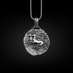 May include: Silver pendant necklace featuring a detailed depiction of a chariot race with two horses and a charioteer. The pendant is round and has a textured surface.