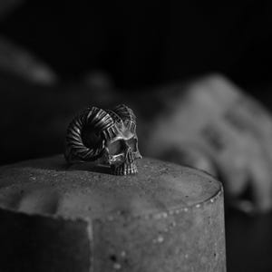 Może przedstawiać: Srebrny pierścionek z czaszką i rogami, pierścionek leży na szarej powierzchni.