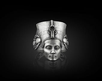 May include: A silver ring featuring a detailed sculpture of an Egyptian queen wearing a crown and headdress.