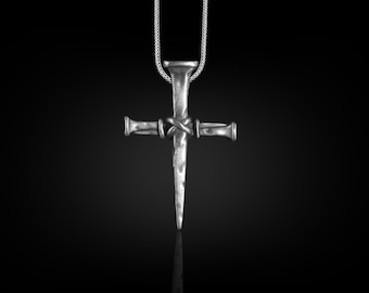 May include: A silver-colored pendant necklace featuring a cross design. The cross is crafted from a nail-like shape with a textured surface and a knot detail at the center. The necklace hangs on a delicate chain against a dark background.