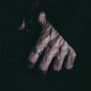 K&ouml;nnte beinhalten: Nahaufnahme einer Hand, die einen silbernen Ring mit einem strukturierten Design tr&auml;gt. Die Hand befindet sich vor einem dunklen Hintergrund.