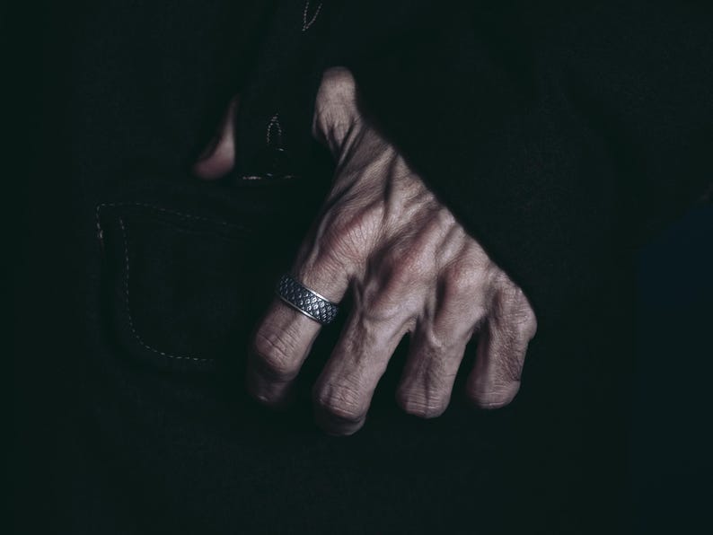 K&ouml;nnte beinhalten: Nahaufnahme einer Hand, die einen silbernen Ring mit einem strukturierten Design tr&auml;gt. Die Hand befindet sich vor einem dunklen Hintergrund.