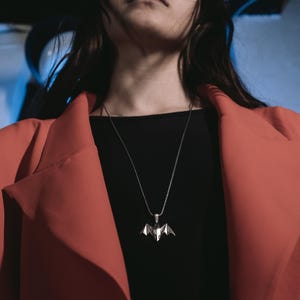 May include: A person wearing a red blazer over a black shirt with a silver bat pendant necklace.