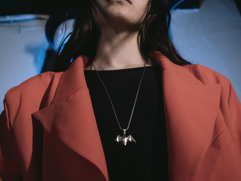 May include: A person wearing a red blazer over a black shirt with a silver bat pendant necklace.
