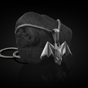 May include: A silver bat pendant necklace with a black rope chain. The pendant is a detailed origami-style bat with wings spread out.