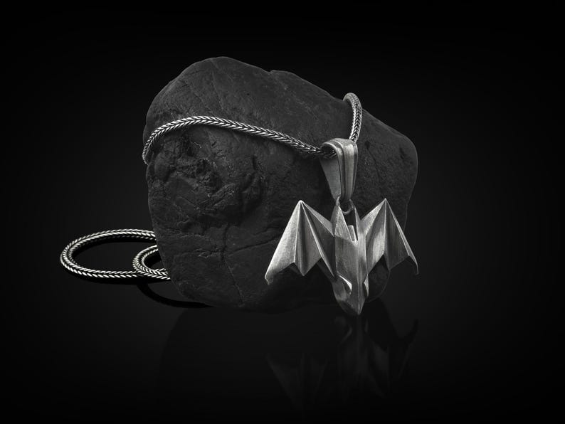 May include: A silver bat pendant necklace with a black rope chain. The pendant is a detailed origami-style bat with wings spread out.