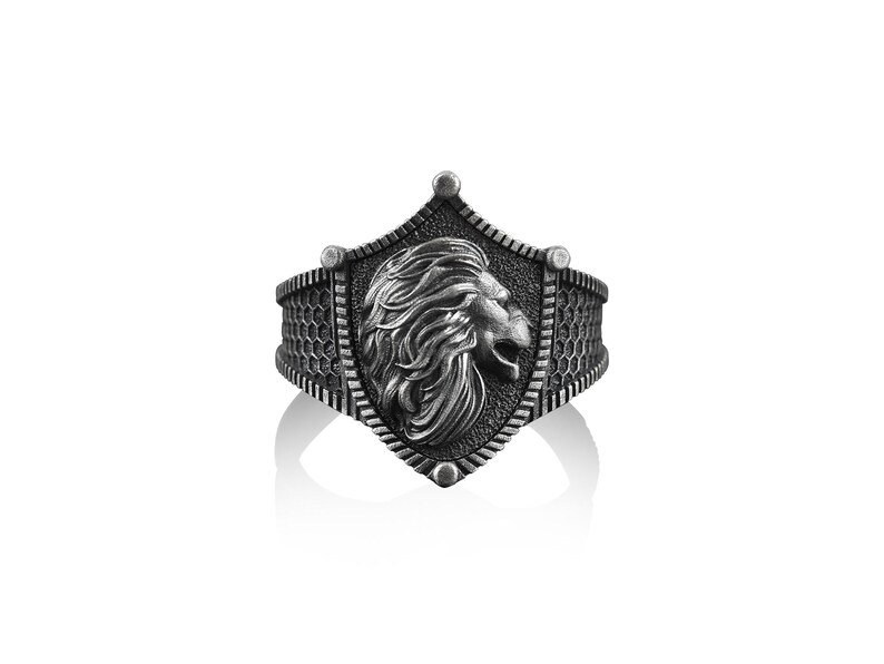 May include: A silver ring with a shield design featuring a lion's head in profile. The shield is surrounded by a textured band.