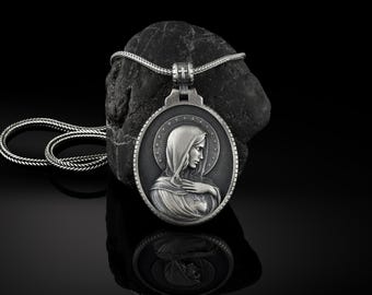 Puede incluir: Un collar de plata con un colgante que muestra una imagen detallada de la Virgen María. El colgante es de forma ovalada y tiene una cadena de plata.