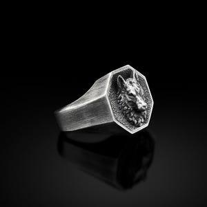 May include: A silver ring with a detailed wolf head design on the face of the ring. The ring is octagonal in shape.
