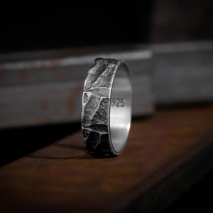 Puede incluir: Un anillo de plata texturizado con una apariencia rugosa, similar a la piedra. El anillo tiene un acabado oscuro y desgastado y el número "925" es visible en el interior. El anillo se muestra sobre una superficie de madera.