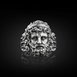 May include: A silver ring with a detailed design of a man's face with a beard and crown. The ring is on a black background.