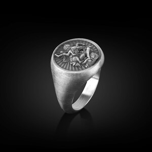 Puede incluir: Un anillo de plata con un dise&ntilde;o detallado de una figura alada sosteniendo una espada. El anillo tiene un acabado cepillado y una forma ovalada redondeada.
