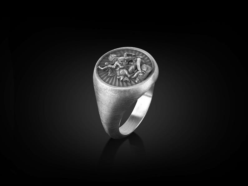 Puede incluir: Un anillo de plata con un dise&ntilde;o detallado de una figura alada sosteniendo una espada. El anillo tiene un acabado cepillado y una forma ovalada redondeada.