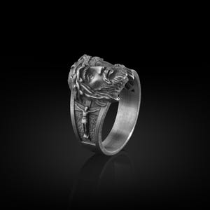May include: A silver ring with a detailed design of a man's face with a crown of thorns and a figure of a man on a cross.