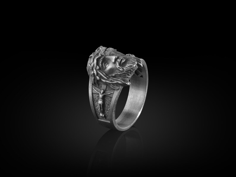 May include: A silver ring with a detailed design of a man's face with a crown of thorns and a figure of a man on a cross.