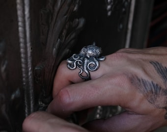 May include: A silver octopus ring with detailed tentacles. The ring is worn on a finger with a tattoo.