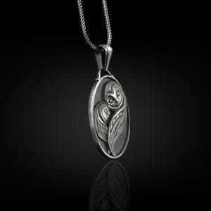 May include: A silver pendant necklace featuring an oval-shaped charm with a detailed, three-dimensional design of an owl. The owl is depicted with its wings folded around its body, and its eyes are visible.