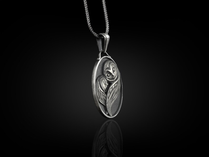 May include: A silver pendant necklace featuring an oval-shaped charm with a detailed, three-dimensional design of an owl. The owl is depicted with its wings folded around its body, and its eyes are visible.