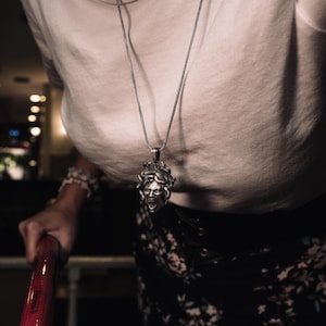 May include: A silver chain necklace with a detailed pendant of a woman's face with snakes for hair. The pendant is worn by a person wearing a white shirt and a floral skirt.