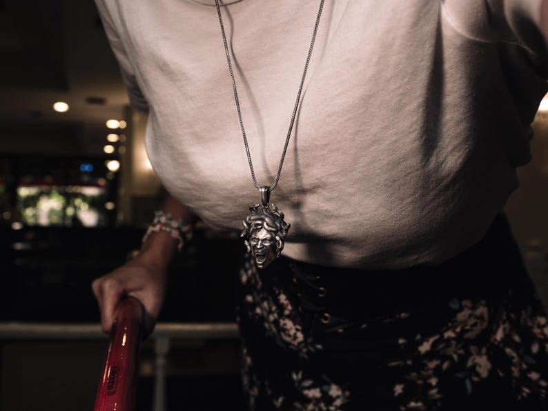 May include: A silver chain necklace with a detailed pendant of a woman's face with snakes for hair. The pendant is worn by a person wearing a white shirt and a floral skirt.