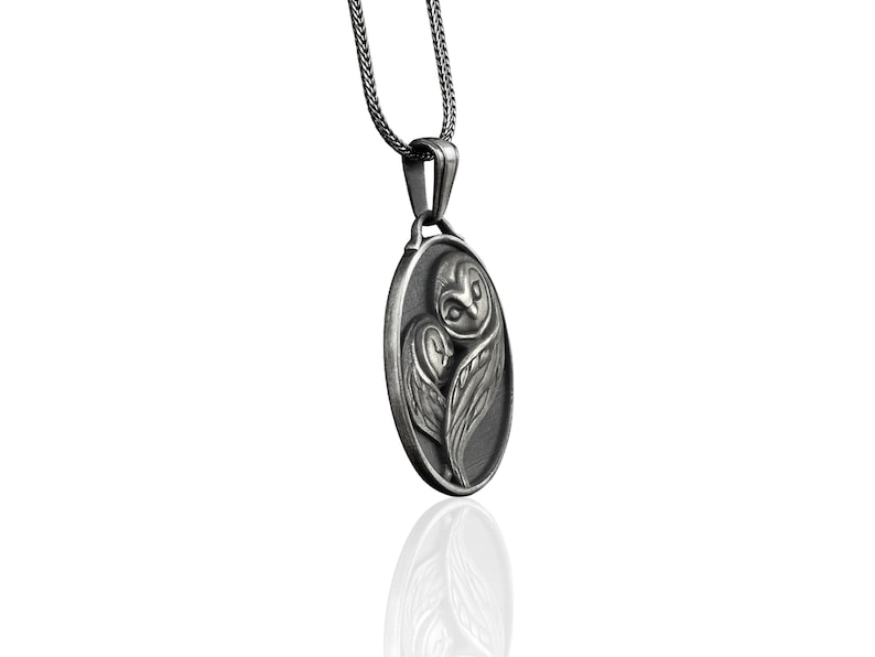 May include: Silver owl pendant necklace with a detailed design of an owl with its wings spread. The pendant is oval shaped and hangs from a silver chain.