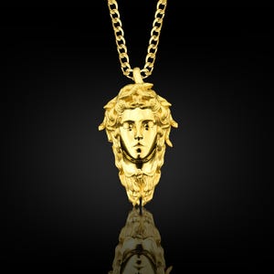 May include: A gold chain necklace with a pendant featuring a detailed, three-dimensional sculpture of a woman's face with a crown of leaves.