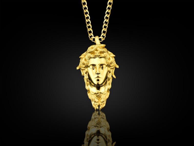 May include: A gold chain necklace with a pendant featuring a detailed, three-dimensional sculpture of a woman's face with a crown of leaves.