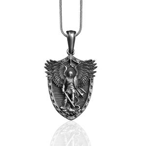 Puede incluir: Collar con colgante de plata con un escudo con un &aacute;ngel que sostiene una espada y una serpiente. El &aacute;ngel tiene alas y est&aacute; representado en una postura protectora.