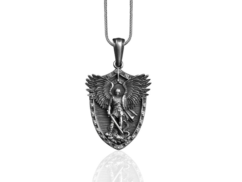 Puede incluir: Collar con colgante de plata con un escudo con un &aacute;ngel que sostiene una espada y una serpiente. El &aacute;ngel tiene alas y est&aacute; representado en una postura protectora.