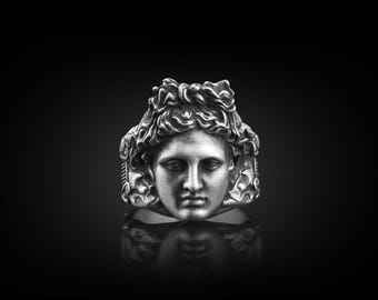 Op de afbeelding: Een zilveren ring met een gedetailleerd beeldhouwwerk van een gezicht met een kroon van haar.