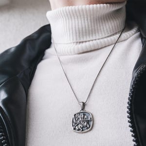 May include: A silver chain necklace with a square pendant featuring a detailed design of a knight on horseback. The necklace is worn over a white turtleneck sweater and a black leather jacket.