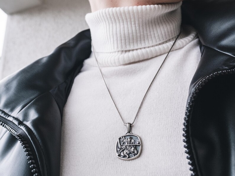 May include: A silver chain necklace with a square pendant featuring a detailed design of a knight on horseback. The necklace is worn over a white turtleneck sweater and a black leather jacket.