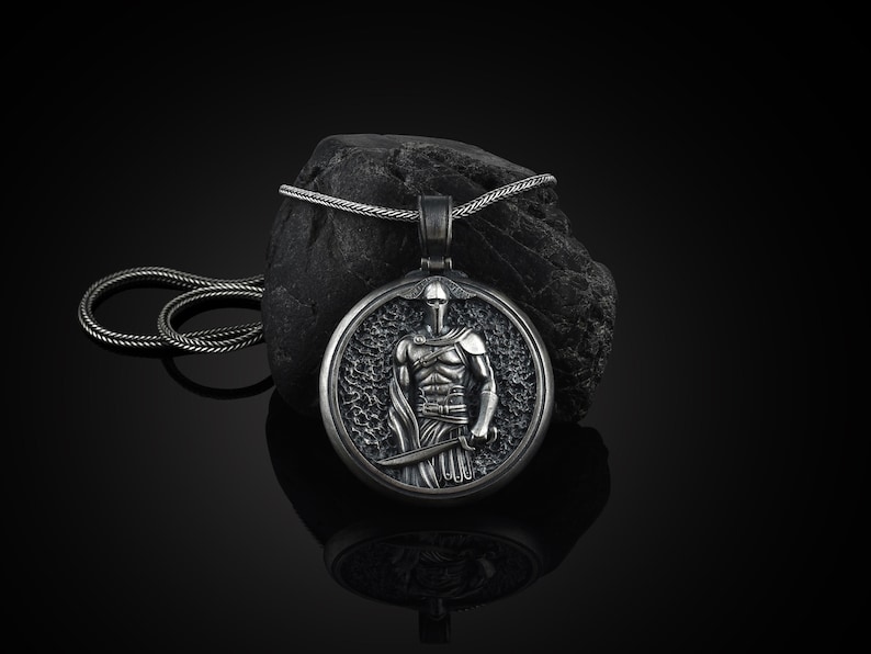 May include: A silver-colored pendant necklace featuring a detailed depiction of a warrior holding a sword. The pendant is circular and set against a dark, textured background. The chain is also silver and lies coiled nearby.