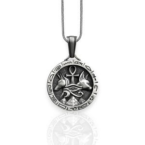 Pu&ograve; includere: Collana con ciondolo in argento con un designo di ispirazione egizia con l'occhio di Horus, il simbolo Ankh e due teste di sciacallo. Il ciondolo &egrave; circondato da un cerchio di geroglifici.