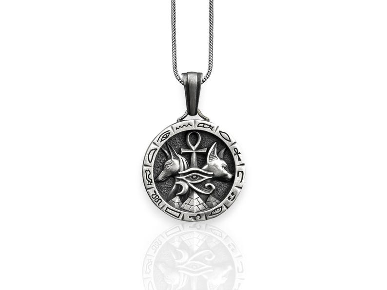 Pu&ograve; includere: Collana con ciondolo in argento con un designo di ispirazione egizia con l'occhio di Horus, il simbolo Ankh e due teste di sciacallo. Il ciondolo &egrave; circondato da un cerchio di geroglifici.