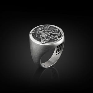 May include: A silver ring with an oval-shaped face featuring a detailed design of an angel with wings spread. The ring has a textured band with a decorative pattern.