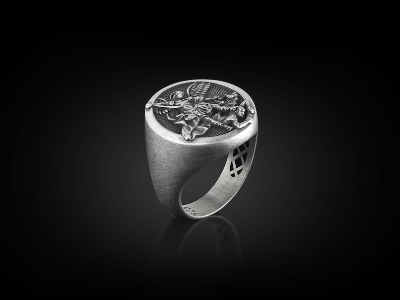May include: A silver ring with an oval-shaped face featuring a detailed design of an angel with wings spread. The ring has a textured band with a decorative pattern.