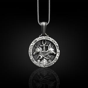 Pu&ograve; includere: Collana con ciondolo in argento di design egizio. Il ciondolo &egrave; un cerchio con un ankh, l'occhio di Horus e due teste di sciacallo. Il cerchio &egrave; circondato da geroglifici.